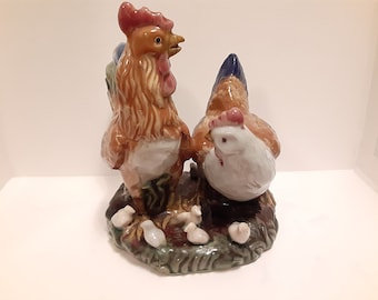 May include: A ceramic rooster and hen figurine set with a brown, white, and blue color scheme. The rooster is standing upright with its head turned to the side. The hen is sitting down with its head turned to the side. There are several small chicks surrounding the rooster and hen.