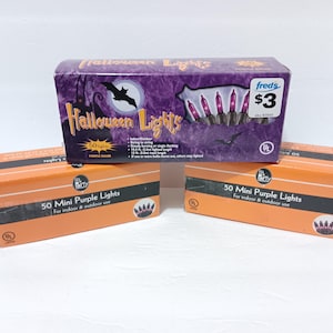 May include: Three boxes of 50 mini purple Halloween lights. One purple box features a bat graphic and text reading Halloween Lights. Two orange boxes are labeled 50 Mini Purple Lights for indoor and outdoor use. Each box displays a UL certification.