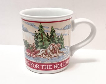 May include: White ceramic mug with a wraparound winter scene featuring a horse-drawn sleigh, evergreen trees, and a cloudy sky. The mug has a red band at the top and bottom with the text "HOME FOR THE HOLIDAY".