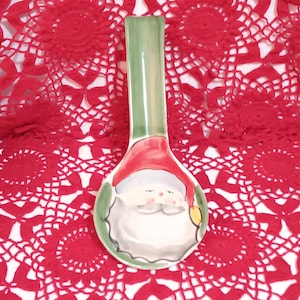 May include: A ceramic spoon rest with a green handle and a Santa Claus face design. The spoon rest has a red hat and a white beard. The background is a red lace tablecloth.