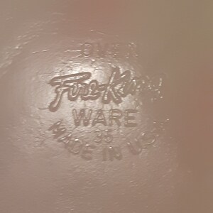 May include: Close-up of a light pink surface with the embossed text "Fire-King WARE 35 MADE IN USA". The text is in a stylized font, indicating a vintage or retro style.