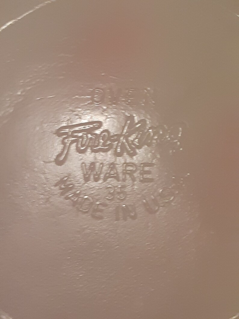 May include: Close-up of a light pink surface with the embossed text "Fire-King WARE 35 MADE IN USA". The text is in a stylized font, indicating a vintage or retro style.