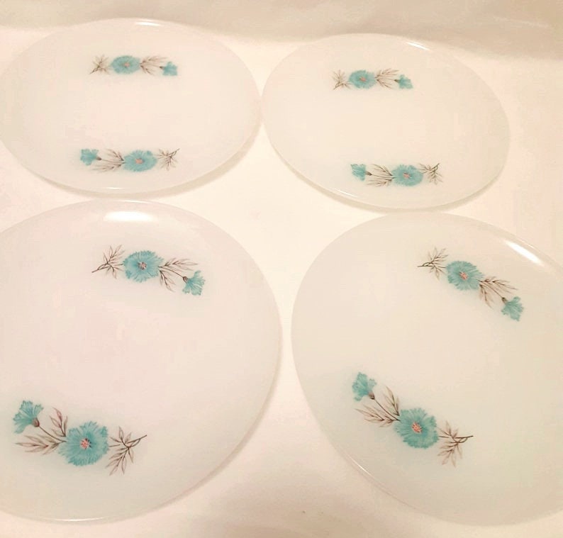 May include: Four white oval plates with a floral design. Each plate features a cluster of turquoise flowers with gray and brown leaves. The plates are arranged in a square formation.