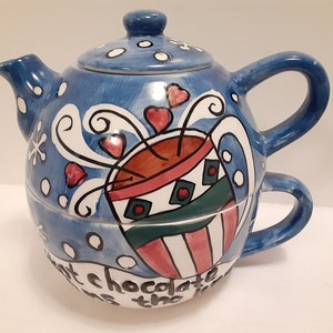 May include: A blue and white ceramic teapot with a mug attached. The teapot has a whimsical design featuring a cup of hot chocolate with steam and hearts. The text on the bottom reads "Hot chocolate is the best".