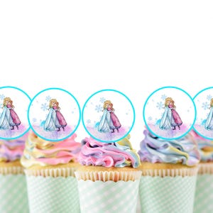 Könnte beinhalten: Sechs Cupcakes, verziert mit blauem und rosa Frosting und gekrönt mit runden, blau umrandeten Toppern, die eine Aquarell-Illustration von zwei Mädchen in Winterkleidung zeigen.
