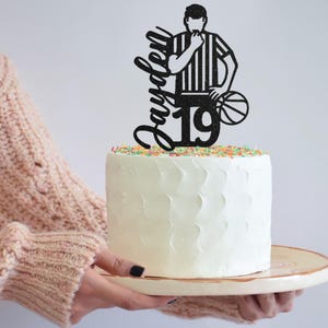 May include: Black glitter cake topper featuring a basketball referee silhouette with the name "Jayden" and the number "19" on a white cake.
