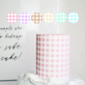 Può includere: Una torta gingham rosa e bianca con un nastro bianco attorno alla base. Ci sono cinque cerchi gingham di diversi colori sopra la torta: rosa, viola, marrone, giallo e verde.
