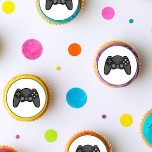 May include: A set of six cupcakes decorated with colourful frosting and edible images of video game controllers. The cupcakes are arranged on a white surface with colourful confetti.