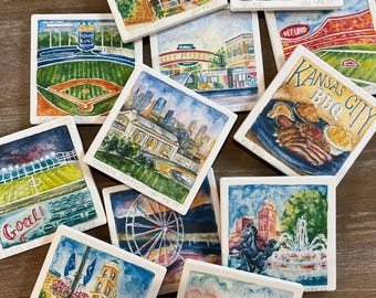 May include: A collection of square ceramic tiles, each hand-painted with watercolor scenes of Kansas City landmarks and attractions. The tiles feature images of baseball stadiums, cityscapes, and the Kansas City BBQ. The tiles are arranged on a wooden surface.