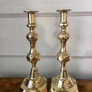 May include: Two brass candlesticks with a hexagonal base and geometric design. The candlesticks have a polished finish and a classic, elegant appearance. The candle holders are approximately 25 cm tall.
