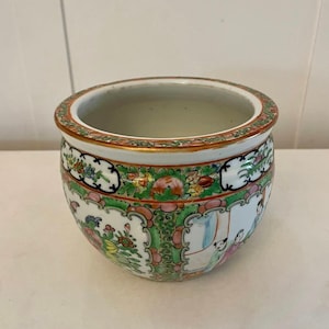 Könnte beinhalten: Ein weißer Porzellantopf mit rundem Körper und breitem Rand. Der Topf ist mit einem bunten Blumenmuster und einer Szene von Menschen in traditioneller chinesischer Kleidung verziert.