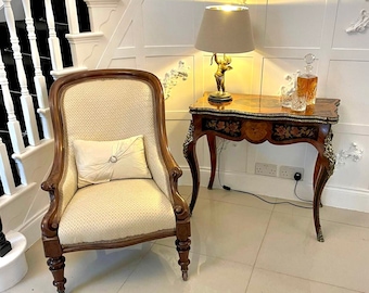 May include: A white armchair with a light brown wooden frame and a white pillow sits next to a small table with a lamp and a decanter. The table has a marquetry top with floral designs and a gold-colored base.