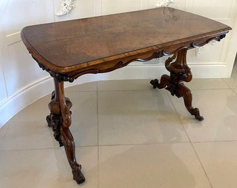 May include: A dark wood table with a burl wood top and intricate carved legs. The table has a curved, scalloped edge and sits on a tiled floor.