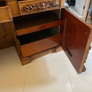 May include: A wooden cabinet with a carved drawer and open shelves. The cabinet has a dark wood finish and is decorated with intricate carvings. The cabinet is open and the shelves are empty.