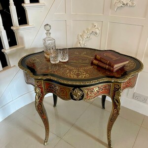 Puede incluir: Una mesa peque&ntilde;a de forma ovalada con incrustaciones de marqueter&iacute;a intrincada en tonos rojos, dorados y negros. La mesa tiene patas ornamentadas y curvadas y un ribete de lat&oacute;n decorativo. Dos libros y una jarra con vasos est&aacute;n sobre la mesa.