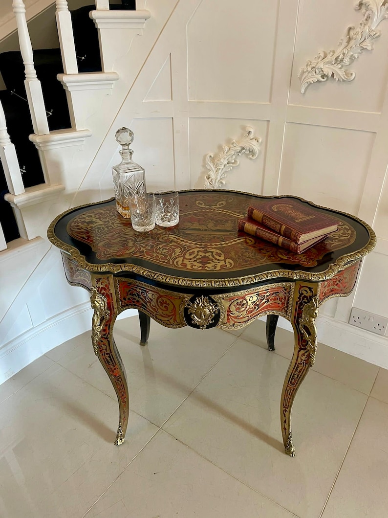 Puede incluir: Una mesa peque&ntilde;a de forma ovalada con incrustaciones de marqueter&iacute;a intrincada en tonos rojos, dorados y negros. La mesa tiene patas ornamentadas y curvadas y un ribete de lat&oacute;n decorativo. Dos libros y una jarra con vasos est&aacute;n sobre la mesa.