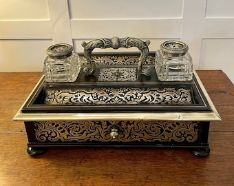 May include: A black wooden desk organizer with a silver trim and a gold and black patterned design. The organizer has two glass inkwells and a drawer with a silver knob.