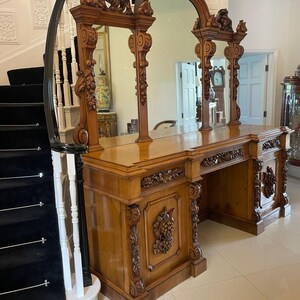 May include: A large, ornately carved wooden vanity with three mirrors. The vanity has a dark wood finish and intricate carvings of leaves and flowers. The mirrors are framed in wood and have a beveled edge.