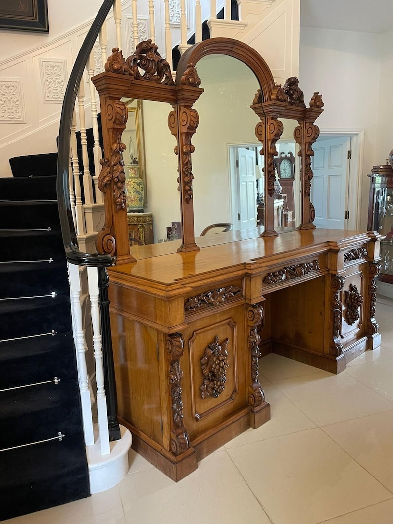 May include: A large, ornately carved wooden vanity with three mirrors. The vanity has a dark wood finish and intricate carvings of leaves and flowers. The mirrors are framed in wood and have a beveled edge.
