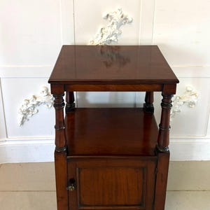 Könnte beinhalten: Ein antiker Beistelltisch aus dunkelbraunem Holz mit quadratischer Oberseite und unterem Regal. Der Tisch hat einen kleinen Schrank mit einer einzigen Tür und dekorativen gedrehten Beinen. Der Tisch ist etwa 61 cm hoch.