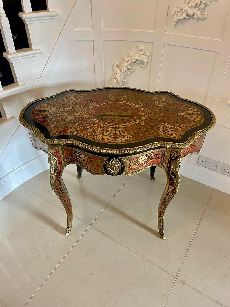 Puede incluir: Una peque&ntilde;a mesa ovalada antigua con incrustaciones de marqueter&iacute;a intrincada en la parte superior. La mesa tiene una base de madera oscura con detalles de lat&oacute;n ornamentados y patas curvas.