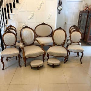 May include: A set of antique furniture, including a loveseat, six chairs, and two footstools. The chairs and loveseat have light-colored patterned upholstery and dark wood frames. The footstools match the chairs. The furniture is arranged in a room with a staircase.