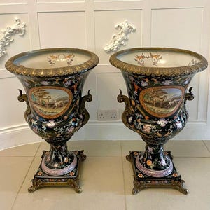 Pode incluir: Um par de grandes urnas de cerâmica preta com bordas e bases de metal em tom de bronze. Cada urna exibe um medalhão oval central com uma cena pintada de um cavalo e um cão, rodeado por padrões florais intrincados e detalhes dourados.