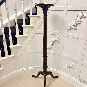 May include: A tall, dark brown wooden plant stand with a round top and ornate carvings. The stand has a slender, fluted column and three curved legs. The stand is approximately 48 inches tall.