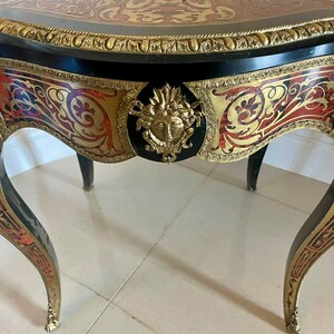 Puede incluir: Una mesa negra y dorada con intrincados detalles de marqueter&iacute;a y una cara tallada en oro en la parte delantera. La mesa tiene una parte superior redondeada y tres patas.