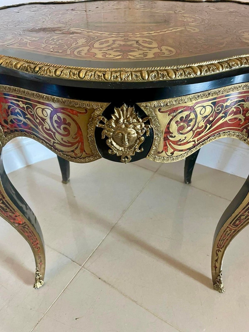 Puede incluir: Una mesa negra y dorada con intrincados detalles de marqueter&iacute;a y una cara tallada en oro en la parte delantera. La mesa tiene una parte superior redondeada y tres patas.
