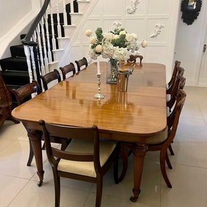 Puede incluir: Una gran mesa de comedor ovalada de madera con un acabado pulido. La mesa está puesta con sillas y un centro de mesa de flores blancas en un jarrón. Una botella de champán y una nevera están sobre la mesa.