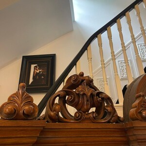 May include: A large, ornate wooden mirror with intricate carvings around the frame. The mirror is reflecting a room with a staircase and a framed picture.