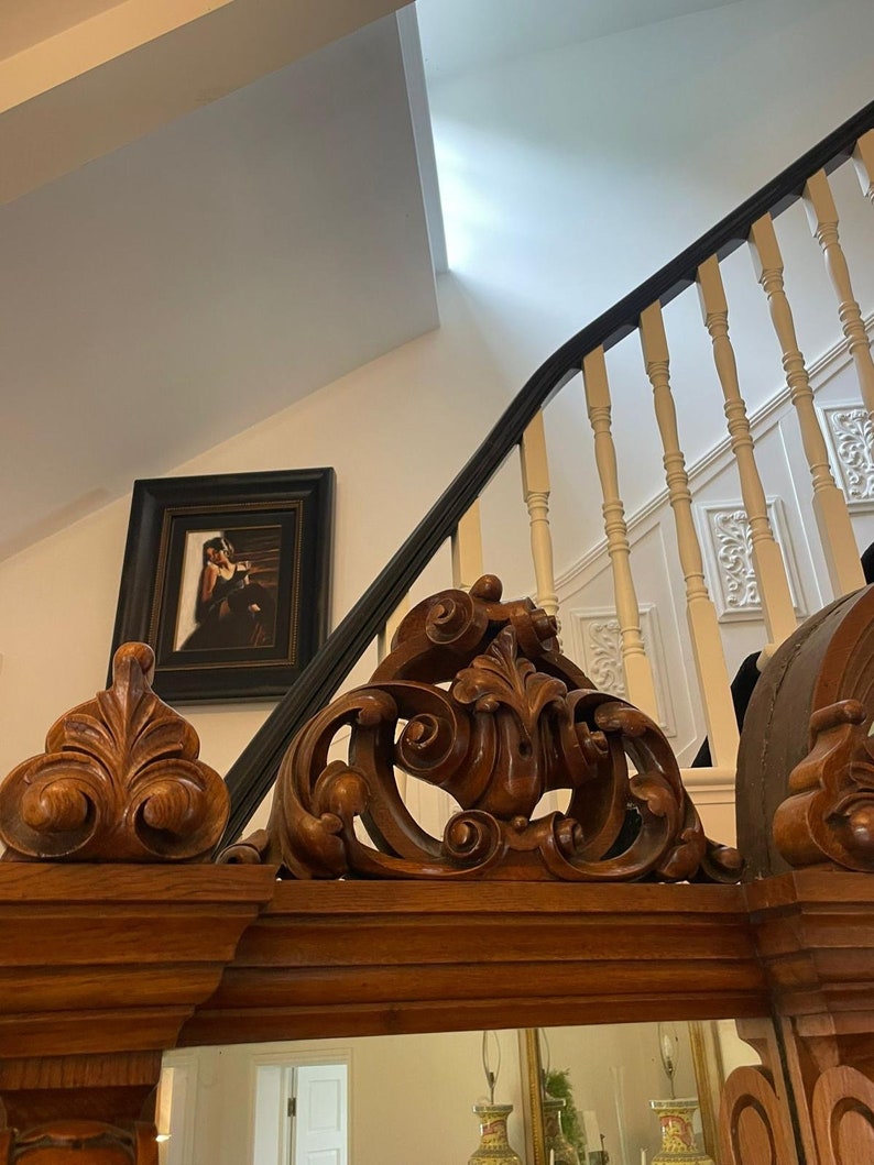 May include: A large, ornate wooden mirror with intricate carvings around the frame. The mirror is reflecting a room with a staircase and a framed picture.