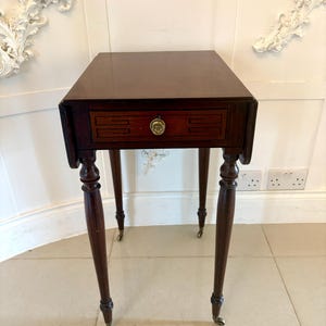 Puede incluir: Una mesa auxiliar antigua de color marrón oscuro con un solo cajón y herrajes de latón. La mesa tiene cuatro patas delgadas y torneadas con pequeñas ruedas. La tapa de la mesa es rectangular. La mesa mide unos 76 cm de altura.