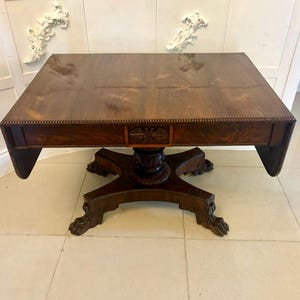 Könnte beinhalten: Ein antiker Holztisch in Dunkelbraun mit quadratischer Tischplatte und kunstvollen Schnitzereien. Der Tisch hat einen Mittelfuß mit vier Klauenfüßen. Der Tisch ist ca. 91 cm breit und 91 cm lang.