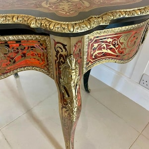 Puede incluir: Una mesa peque&ntilde;a y adornada con un acabado negro y dorado. La superficie de la mesa est&aacute; incrustada con intrincados patrones de rojo, dorado y negro. La mesa tiene tres patas, cada una con un dise&ntilde;o tallado.