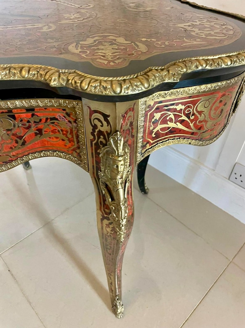 Puede incluir: Una mesa peque&ntilde;a y adornada con un acabado negro y dorado. La superficie de la mesa est&aacute; incrustada con intrincados patrones de rojo, dorado y negro. La mesa tiene tres patas, cada una con un dise&ntilde;o tallado.