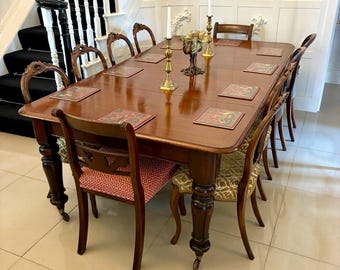 May include: A large, polished wooden dining table with eight matching chairs. The table is rectangular with rounded corners and sits on ornate, turned legs. The chairs have patterned upholstery and curved backs. Gold candlesticks and a decorative bowl adorn the table's center.