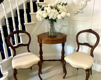 May include: Two antique wooden chairs with white upholstery and a small round wooden table with a white vase of white flowers on top. The chairs and table are in a hallway with a white staircase in the background.