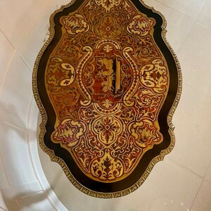 Puede incluir: Una tapa de mesa redonda y ornamentada con fondo negro e incrustaciones de marqueter&iacute;a intrincadas en tonos dorados, rojos y marrones. La tapa de la mesa tiene un borde festoneado y est&aacute; rodeada por un ribete dorado.