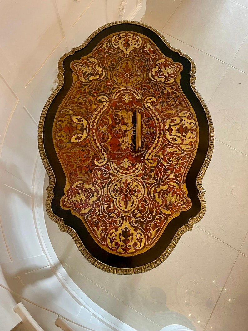 Puede incluir: Una tapa de mesa redonda y ornamentada con fondo negro e incrustaciones de marqueter&iacute;a intrincadas en tonos dorados, rojos y marrones. La tapa de la mesa tiene un borde festoneado y est&aacute; rodeada por un ribete dorado.