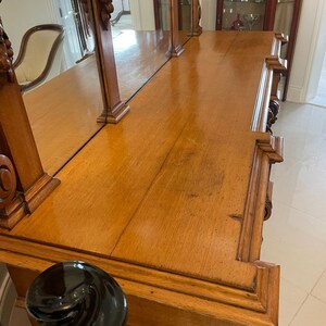 May include: A wooden antique dresser with a mirrored top and intricate carvings. The dresser has a dark wood finish and a black base.
