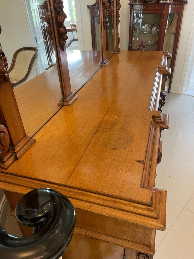 May include: A wooden antique dresser with a mirrored top and intricate carvings. The dresser has a dark wood finish and a black base.