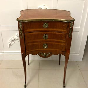 May include: A small, antique wooden chest of drawers with three drawers. The chest has a curved front, brass hardware, and slender, curved legs. The top is a lighter shade of brown than the rest of the chest. The chest is approximately 30 inches tall.