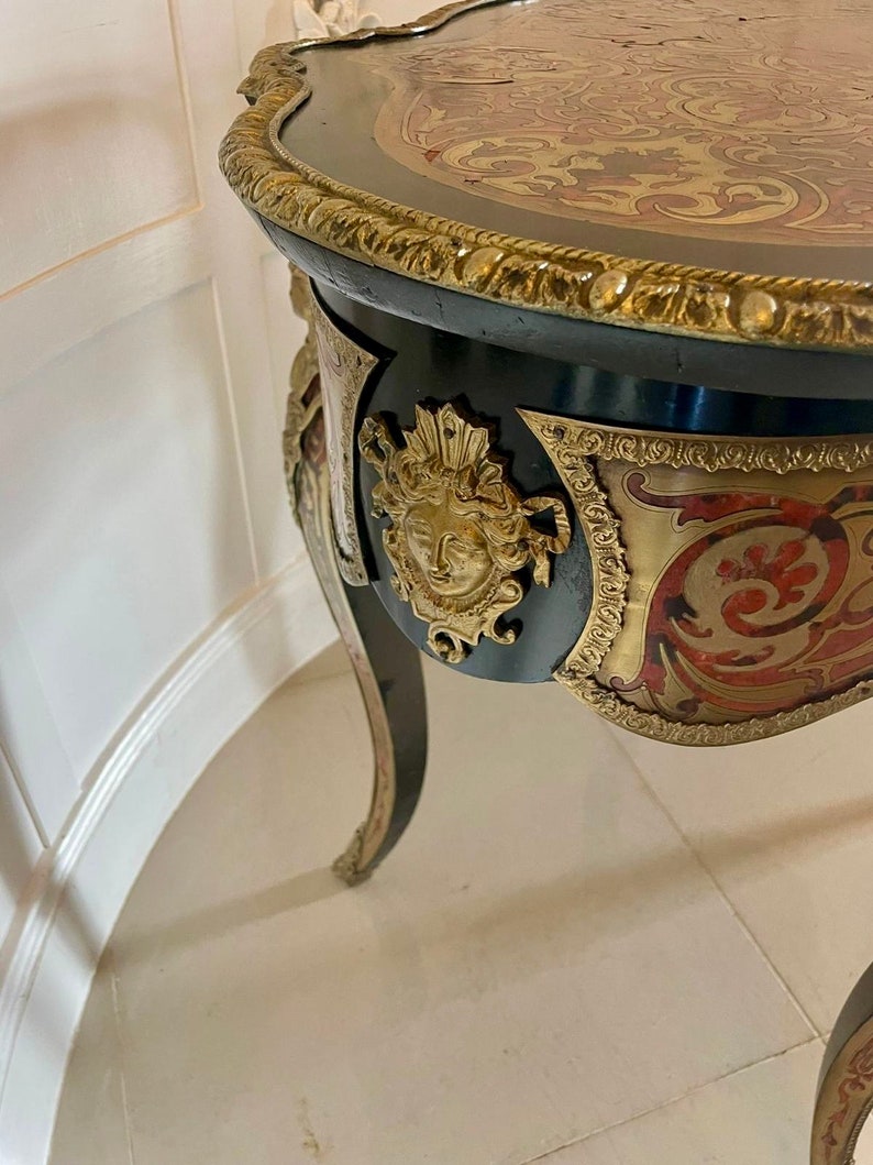 Puede incluir: Una mesa ornamentada negra y dorada con una tapa de marqueter&iacute;a y un dise&ntilde;o tallado. La mesa tiene tres patas y un borde decorativo alrededor de la parte superior.