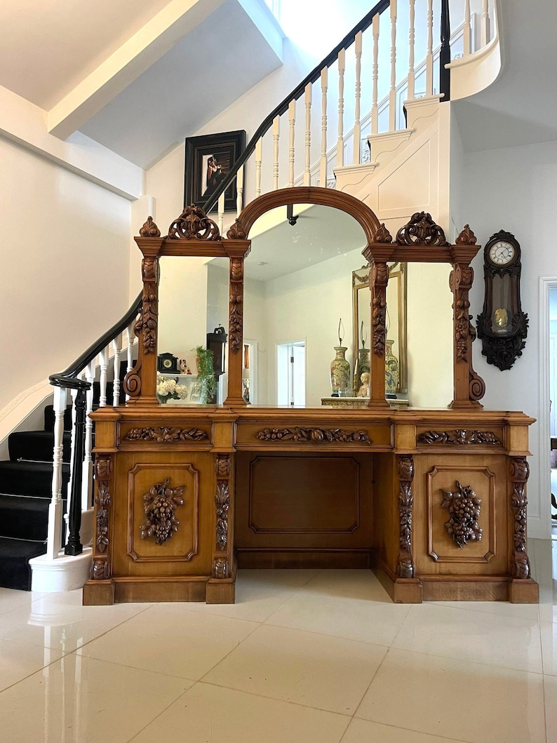 May include: A large, ornately carved wooden sideboard with three mirrors and a central arched mirror. The sideboard has a dark wood finish and intricate carvings of grapes and leaves.