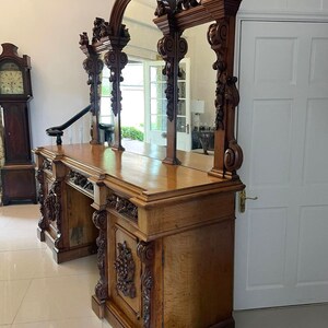May include: A large, ornately carved wooden sideboard with a triple mirror back. The sideboard has a dark wood finish and intricate carvings of flowers and leaves. The mirror is arched and has a beveled edge.