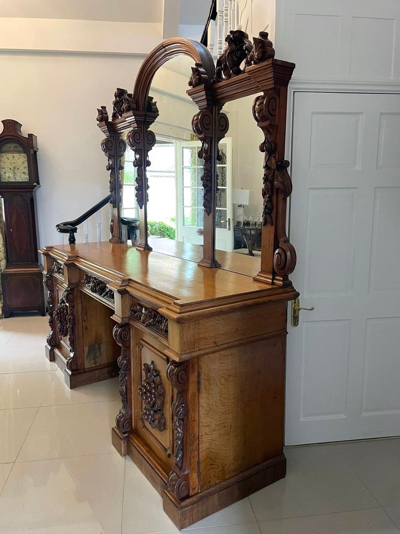 May include: A large, ornately carved wooden sideboard with a triple mirror back. The sideboard has a dark wood finish and intricate carvings of flowers and leaves. The mirror is arched and has a beveled edge.