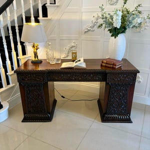 May include: A dark wood desk with ornate carvings, featuring a lamp, books, and a vase of flowers. The desk has a rectangular top and two carved pedestals. A gold-colored lamp with a cherub base sits on the desk.