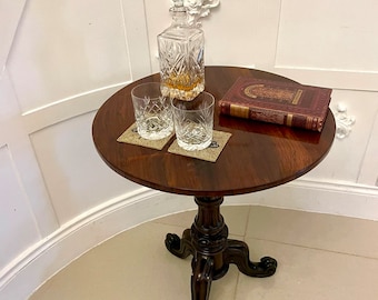 May include: A dark wood round table with a carved base and three legs. On the table are two clear glass tumblers, a crystal decanter with a stopper, and a red leather-bound book.